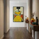 Contemporary figurative painting depicting a woman in a vibrant yellow dress with black hair and gestural brushstrokes, soft cream and ochre palette with red accents, hand-painted canvas in modern hallway