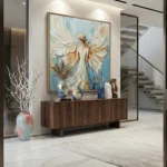 Figurative hand-painted canvas depicting an angel with expansive wings in cream, gold, and soft blue tones, contemporary entryway