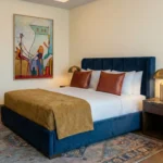 Figurative hand-painted canvas with abstract musicians and architectural elements in warm ochre and teal tones, modern bedroom setting