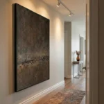 Textured abstract hand-painted canvas in charcoal and bronze tones with weathered surface detail, contemporary hallway